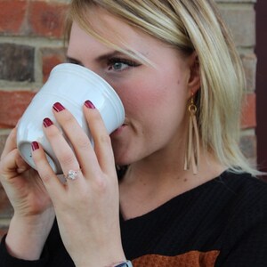 May include: A woman wearing a black and brown sweater with a light blue watch, drinks from a white ceramic mug. She is wearing a ring on her left ring finger and long, dangling earrings.