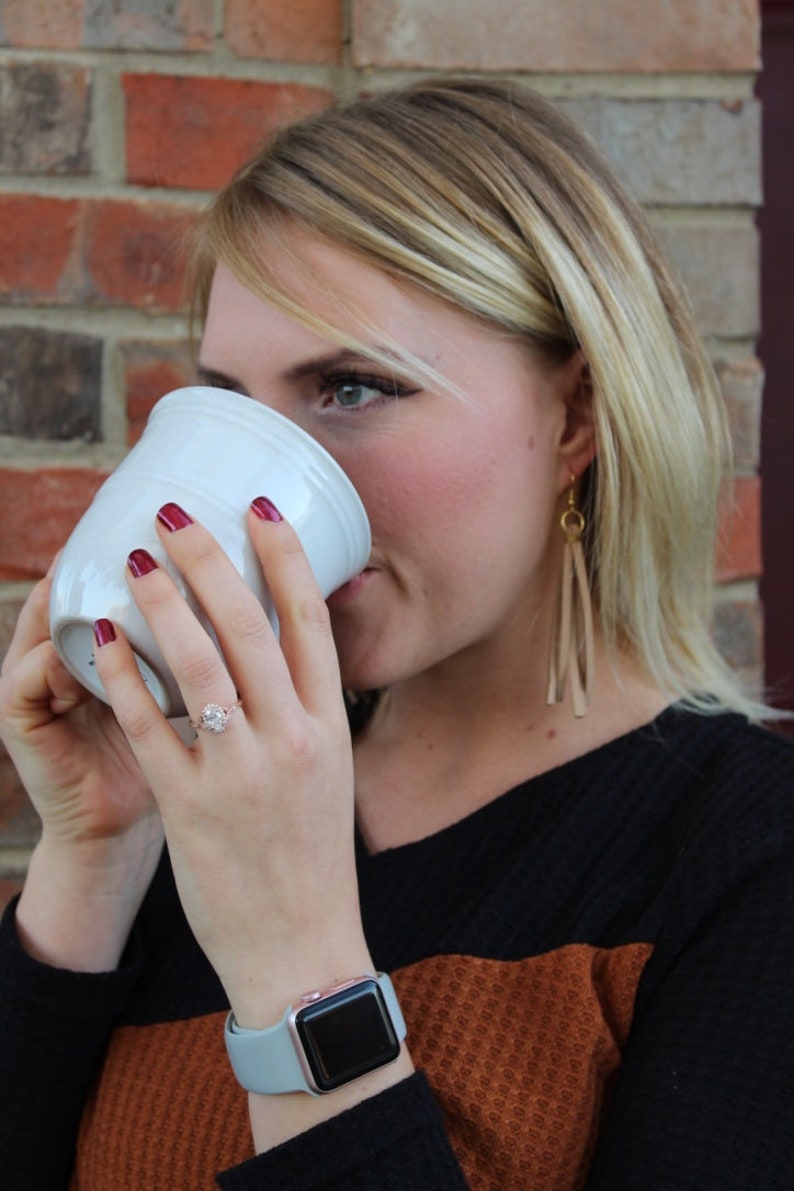 May include: A woman wearing a black and brown sweater with a light blue watch, drinks from a white ceramic mug. She is wearing a ring on her left ring finger and long, dangling earrings.