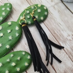 May include: Black suede tassel earrings with gold hoops. The earrings are hanging from a cactus.