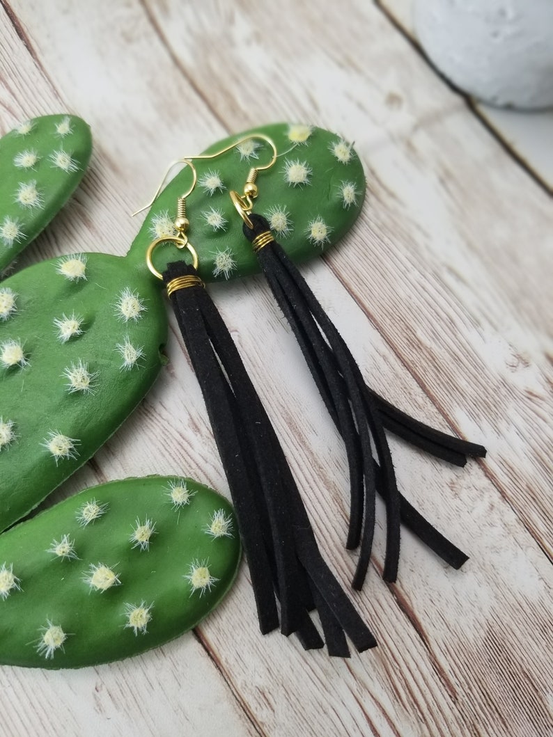 May include: Black suede tassel earrings with gold hoops. The earrings are hanging from a cactus.