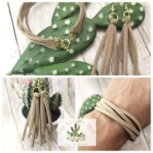 May include: A set of brown suede earrings and a matching bracelet. The earrings are tassel style and the bracelet is a wrap style. The jewelry is displayed on a cactus and a person's wrist.