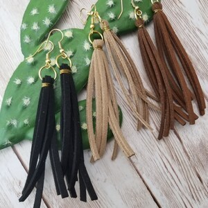 May include: Three pairs of suede tassel earrings in black, brown, and beige. The earrings are hanging from a cactus on a wooden background.