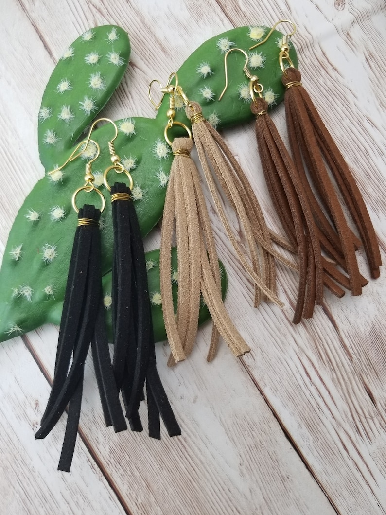 May include: Three pairs of suede tassel earrings in black, brown, and beige. The earrings are hanging from a cactus on a wooden background.