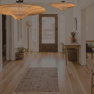 Pode incluir: Um corredor de madeira clara com piso de madeira e um tapete. Duas luzes pendentes de tecido penduradas no teto. Uma mesa de madeira com uma cesta de vime fica encostada na parede. Uma porta leva para o exterior.