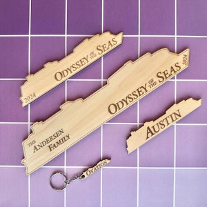 Könnte beinhalten: Holz-Souvenirs in Form von Kreuzfahrtschiffen mit graviertem Text. Die Artikel umfassen einen Schlüsselanhänger, ein Stück mit "Austin" und Stücke mit "Odyssey of the Seas 2024" und "The Andersen Family". Die Artikel sind hellbraun.