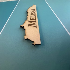 K&ouml;nnte beinhalten: Ein Holz-Ausschnitt eines Kreuzfahrtschiffs mit dem eingravierten Namen "Melissa". Der Ausschnitt ist hellbraun und liegt auf einer blauen Oberfl&auml;che.