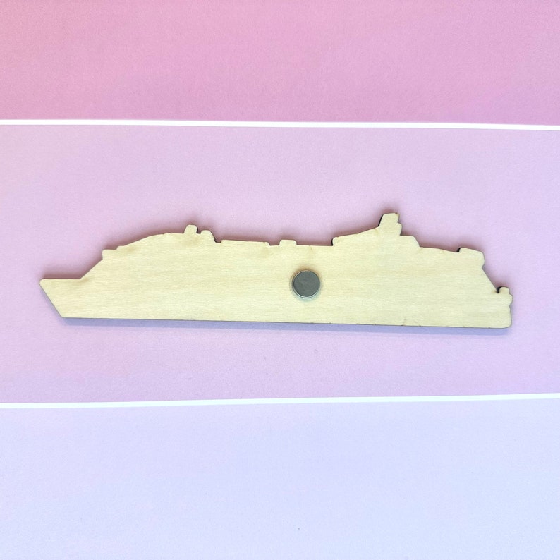 K&ouml;nnte beinhalten: Holzschnitt eines Kreuzfahrtschiffs mit einem Magneten auf der R&uuml;ckseite. Der Ausschnitt ist hellbraun und befindet sich auf einem rosa Hintergrund.