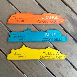 K&ouml;nnte beinhalten: Drei Holz-Schiffs-Umrisse, in Orange, Blau und Gelb bemalt. Jedes Schiff tr&auml;gt den Namen der Reederei und das Baujahr. Das orangefarbene Schiff ist die "Odyssey of the Seas", gebaut im Jahr 2021. Das blaue Schiff ist die "Icon of the Seas", gebaut im Jahr 2024. Das gelbe Schiff ist die "Oasis of the Seas", gebaut im Jahr 2018.