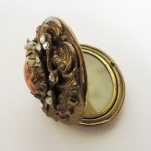 May include: An antique gold-toned locket with an oval shape, featuring ornate scrollwork and small, clear gemstones. The locket opens to reveal a space for a photograph or keepsake. A small painted image is visible on the front.