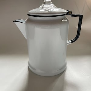 May include: A white enamel coffee pot with a black handle and trim. The pot has a clear glass lid and a spout for pouring. The pot is designed for stovetop use and is a classic kitchen item.