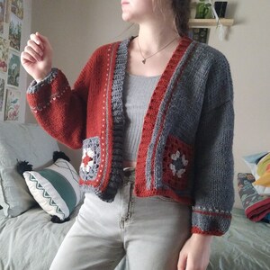 May include: A gray and red knitted cardigan with crocheted details. The cardigan has a V-neck and is open in the front. The cardigan has a crocheted flower design on the front pockets.