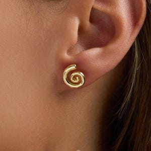 May include: A close-up of a gold spiral stud earring. The earring is a simple, elegant design with a coiled shape. The earring is worn on an ear, and the skin tone is light. The earring is shiny and reflects light.