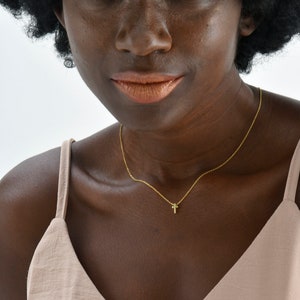 May include: A woman with dark brown skin and a large afro hairstyle is wearing a light brown sleeveless top. She is wearing a gold chain necklace with a small cross pendant.