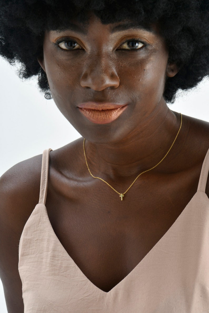 May include: A woman with dark brown skin and a large afro hairstyle is wearing a light brown sleeveless top. She is wearing a gold chain necklace with a small cross pendant.