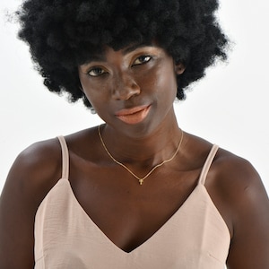 May include: A woman with dark skin and a large afro wig is wearing a light pink sleeveless top and a gold necklace with a small cross pendant.