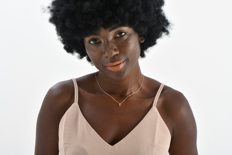 May include: A woman with dark skin and a large afro wig is wearing a light pink sleeveless top and a gold necklace with a small cross pendant.