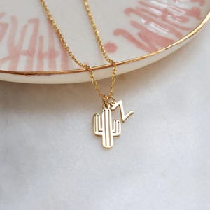 Op de afbeelding: Een goudkleurige ketting met een cactus- en een zigzagbedel. De ketting ligt op een wit oppervlak met een roze en goudkleurige schaal op de achtergrond. De ketting is delicaat en de bedels zijn minimalistisch.