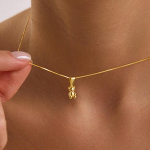 May include: A gold-toned necklace featuring a small, three-dimensional teddy bear pendant suspended from a delicate box chain. The pendant has a polished, reflective surface and is attached to the chain by a simple, matching gold-toned bail.