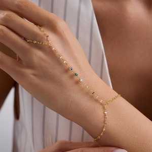May include: A delicate gold hand chain featuring a series of small, colorful gemstones. The chain connects to a bracelet and extends to a ring, creating a unique jewelry piece. The gemstones include purple, green, and clear stones.
