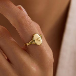 以下が含まれることがあります： 金色のシグネットリング。楕円形の面と装飾的なディテールが特徴です。リングには、筆記体フォントで「Jayna」という名前と小さな星が刻まれています。指にはめられたリングです。