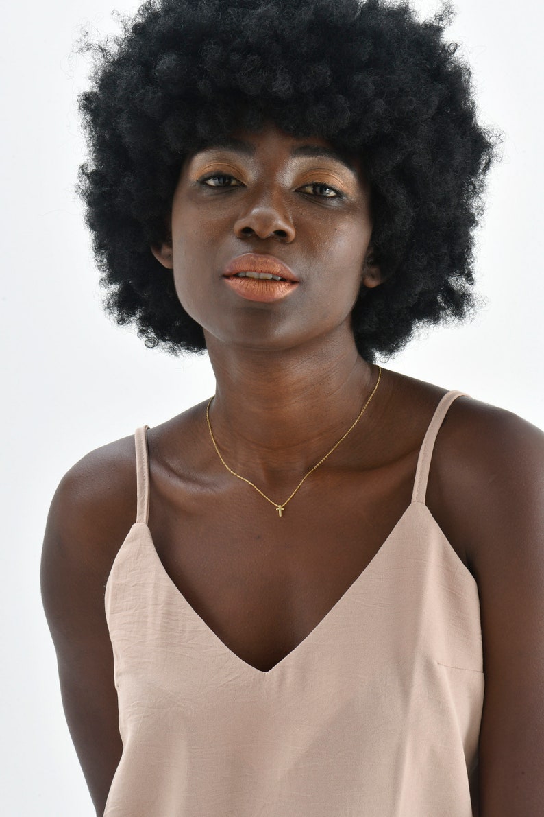 May include: A woman with an afro hairstyle wearing a light brown sleeveless top with a thin gold chain necklace with a small cross pendant.