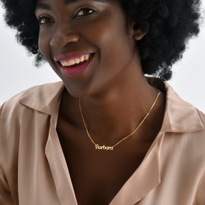 Op de afbeelding: Een vrouw met een afro-kapsel draagt een lichtbruin shirt en een gouden ketting met de naam "Barbara" erop.