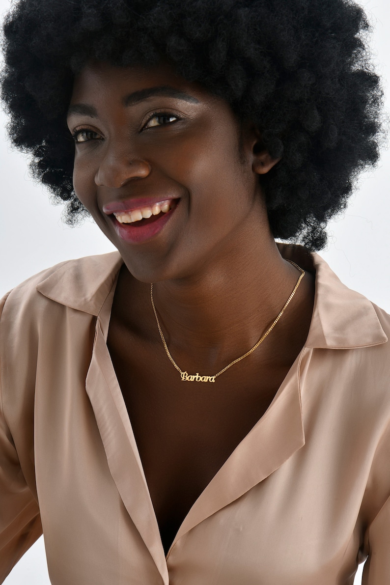 Op de afbeelding: Een vrouw met een afro-kapsel draagt een lichtbruin shirt en een gouden ketting met de naam "Barbara" erop.