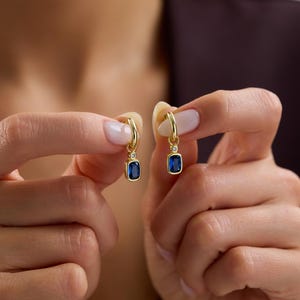 May include: Gold hoop earrings, each with a rectangular, deep blue gemstone and a small, clear accent stone. The earrings are displayed against a neutral background, highlighting their design and colour.