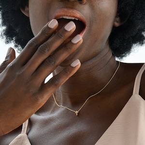 May include: A woman with dark curly hair is wearing a light brown sleeveless top and a gold necklace with a small cross pendant. She is looking up and has her hand covering her mouth.