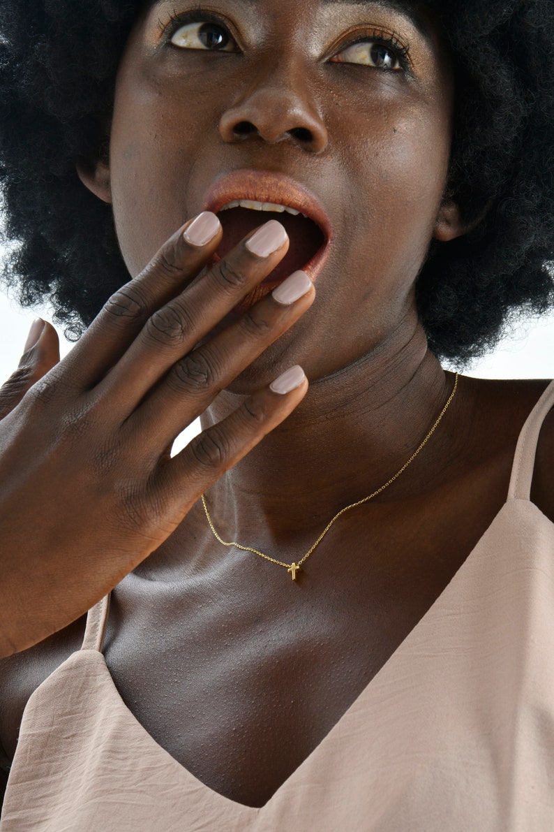 May include: A woman with dark curly hair is wearing a light brown sleeveless top and a gold necklace with a small cross pendant. She is looking up and has her hand covering her mouth.