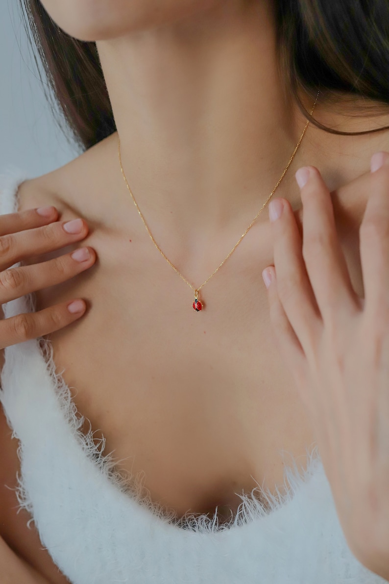 K&ouml;nnte beinhalten: Eine goldene Kettenkette mit einem kleinen roten Edelstein-Anh&auml;nger. Die Kette wird von einer Person mit langen dunklen Haaren getragen.