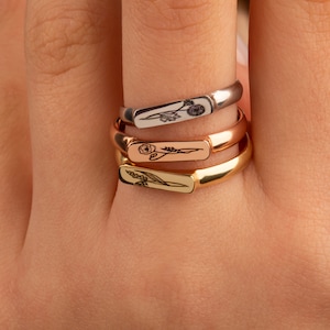 May include: Three gold rings with floral designs engraved on the bands. The rings are stacked on a finger.
