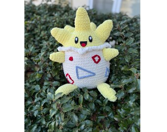 May include: A handmade plush toy in the shape of a Togepi character. The plush features a yellow star-shaped head, small black eyes, and a smiling mouth. The body is white with red and blue geometric shapes.