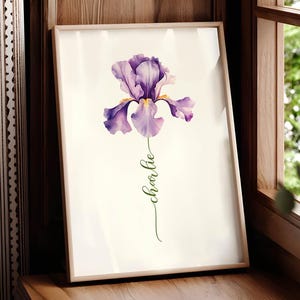 May include: Framed watercolor print featuring a purple iris flower with green stem and the name "charlie" in cursive. The artwork is set against a cream background, framed in a light wood, and displayed on a wooden surface.