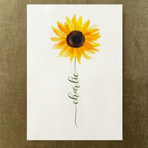 May include: A rectangular white card featuring a watercolor illustration of a single sunflower with bright yellow petals and a dark brown center. A thin green stem extends downward, forming the cursive text Charlie in a flowing script style.