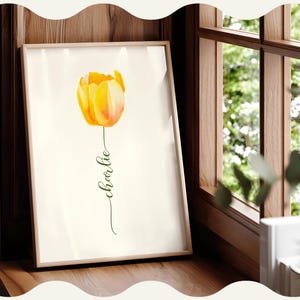 Może przedstawiać: Oprawiony wydruk artystyczny przedstawiający akwarelowy tulipan w odcieniach żółci i pomarańczy. Słowo "Charlie" jest napisane zielonym skryptem pod kwiatem. Grafika jest wyświetlana na drewnianej powierzchni w pobliżu okna.
