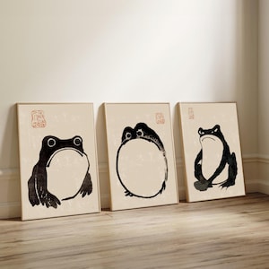 Peut inclure: Trois impressions d'art encadrées représentant des illustrations de grenouilles à l'encre noire sur fond beige. Chaque grenouille a une pose et une expression uniques. Les impressions sont exposées sur un sol en bois, adossées à un mur blanc.