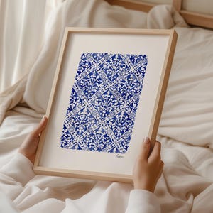 May include: A framed art print featuring a blue and white watercolor pattern of repeating diamond shapes filled with intricate floral motifs. The artwork is set against a white background within a light wood frame and includes the word Lisbon.
