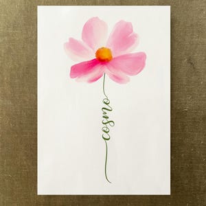 Può includere: Un dipinto ad acquerello su carta bianca raffigurante un singolo fiore di cosmea rosa con un centro arancione brillante. Il gambo del fiore è formato dal testo corsivo verde "cosmo" che si estende verso il basso in uno stile fluido.