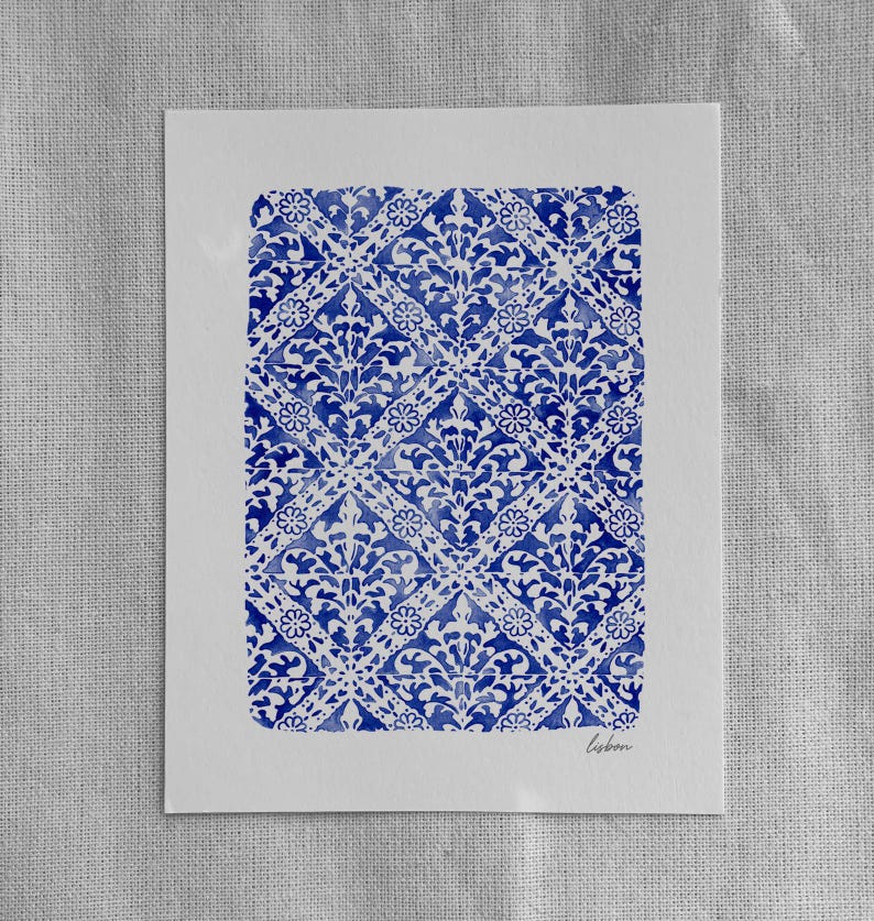 May include: A rectangular white paper print featuring a repeating blue watercolor pattern of diamonds, floral motifs, and intricate scrollwork. The word "lisbon" is written in small, cursive, dark gray script in the bottom right corner of the print.