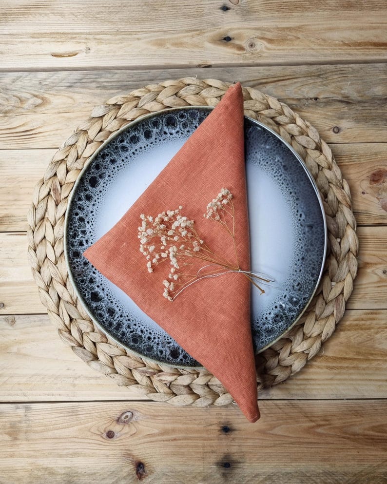 May include: A triangular folded burnt orange linen napkin placed on a round ceramic plate with a white center and a dark blue speckled rim, topped with a small sprig of dried white baby's breath flowers, resting on a woven round placemat.