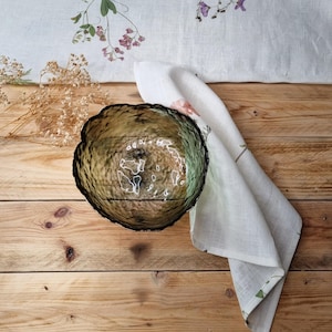 May include: A round, translucent olive green glass bowl with a textured, hammered surface and an irregular, wavy rim. The bowl is positioned next to a white linen napkin featuring a delicate pressed flower pattern and a small bunch of dried baby's breath.