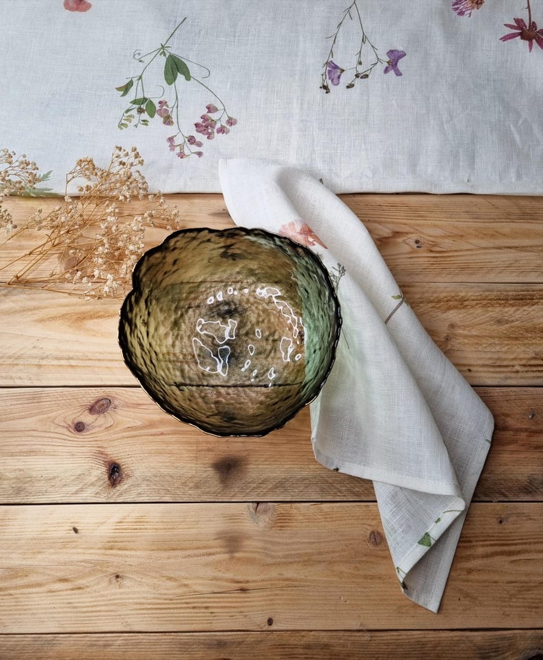 May include: A round, translucent olive green glass bowl with a textured, hammered surface and an irregular, wavy rim. The bowl is positioned next to a white linen napkin featuring a delicate pressed flower pattern and a small bunch of dried baby's breath.