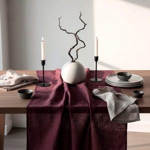 Könnte beinhalten: Ein gedeckter Esstisch mit bordeauxfarbenem Tischläufer, weißen Tellern, kleinen schwarzen Schalen und Leinen-Servietten. Schwarze Kerzenhalter mit weißen Kerzen und eine weiße Vase mit Zweigen vervollständigen das Arrangement.