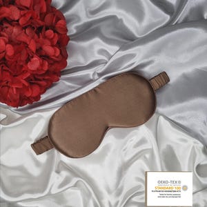 May include: A brown silk sleep mask on a white satin background. The mask has a thin elastic band and is labeled with the Oeko-Tex Standard 100 certification.