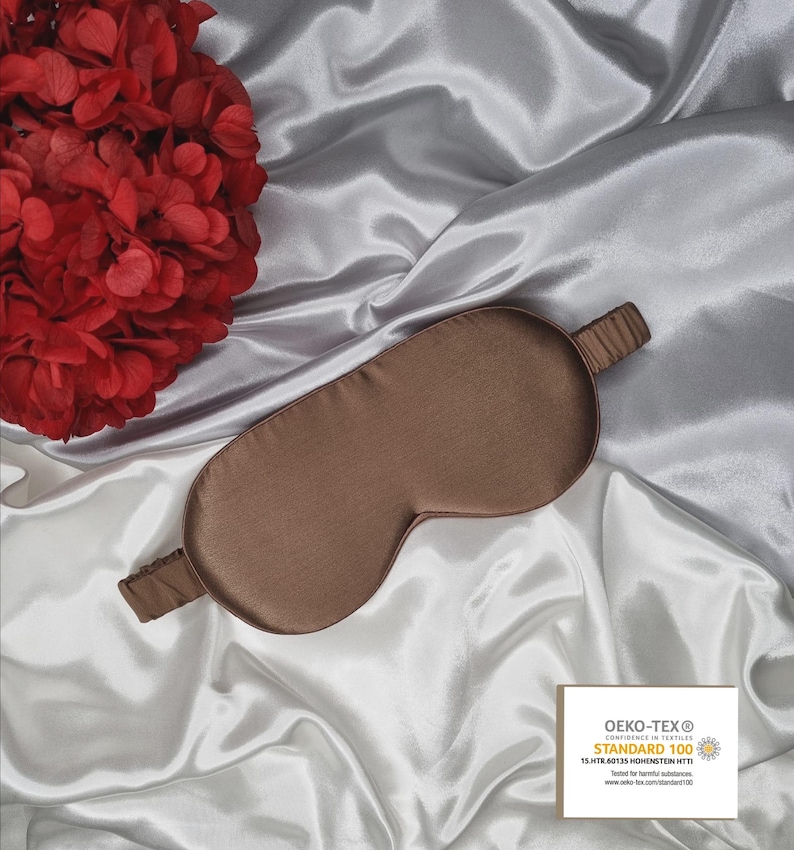 May include: A brown silk sleep mask on a white satin background. The mask has a thin elastic band and is labeled with the Oeko-Tex Standard 100 certification.