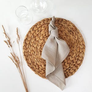 May include: A round, woven brown water hyacinth placemat topped with a light beige linen napkin tied in a simple knot. Beside the placemat are three dried wheat stalks and two clear glass vessels, including a carafe and a small drinking glass.