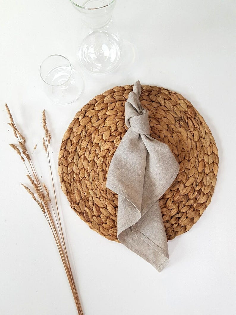 May include: A round, woven brown water hyacinth placemat topped with a light beige linen napkin tied in a simple knot. Beside the placemat are three dried wheat stalks and two clear glass vessels, including a carafe and a small drinking glass.