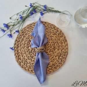 May include: A light blue linen napkin folded through a braided natural fiber napkin ring, placed on a round woven placemat. Beside the placemat are blue cornflowers and a clear glass carafe. The text LeMoor is printed in the bottom right corner.