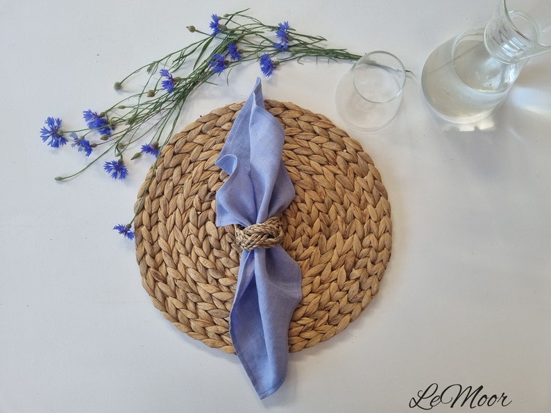 May include: A light blue linen napkin folded through a braided natural fiber napkin ring, placed on a round woven placemat. Beside the placemat are blue cornflowers and a clear glass carafe. The text LeMoor is printed in the bottom right corner.